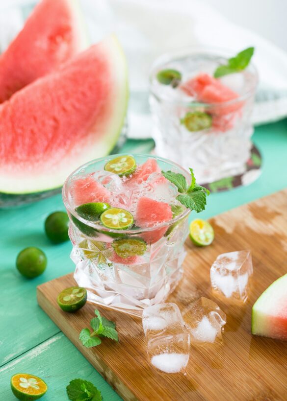 A watermelon and lime cocktail over ice with fresh mint, beside watermelon slices on a green table