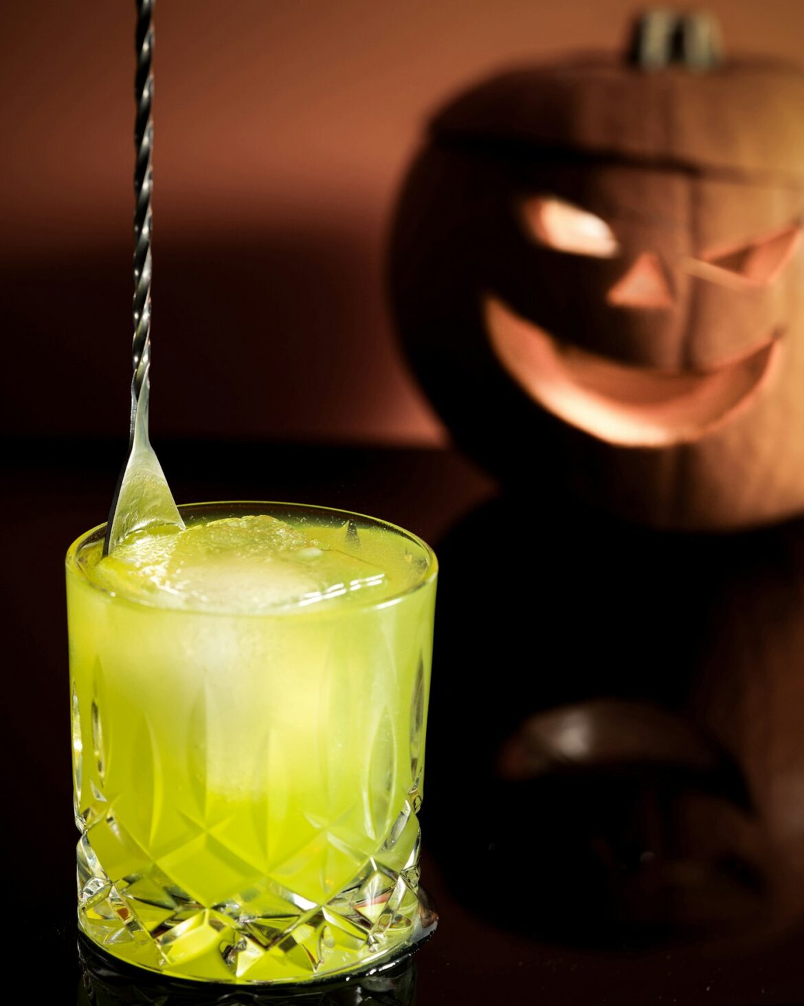Bright green Halloween cocktail in a cut glass tumbler with a lit jack o lantern behind it