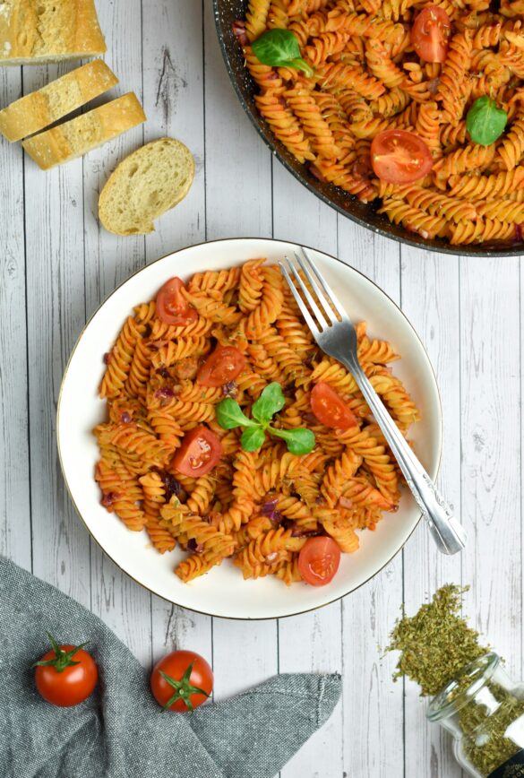 A plate of fusilli pasta in tomato sauce with cherry tomatoes and basil, beside the skillet and crusty bread