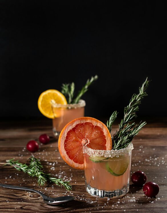 Two autumn cocktails with sugared rims, grapefruit slices, rosemary sprigs and fresh cranberries on a dark wooden table