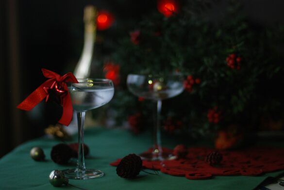 Two coupe cocktail glasses tied with a red ribbon beside a bottle and a decorated Christmas tree