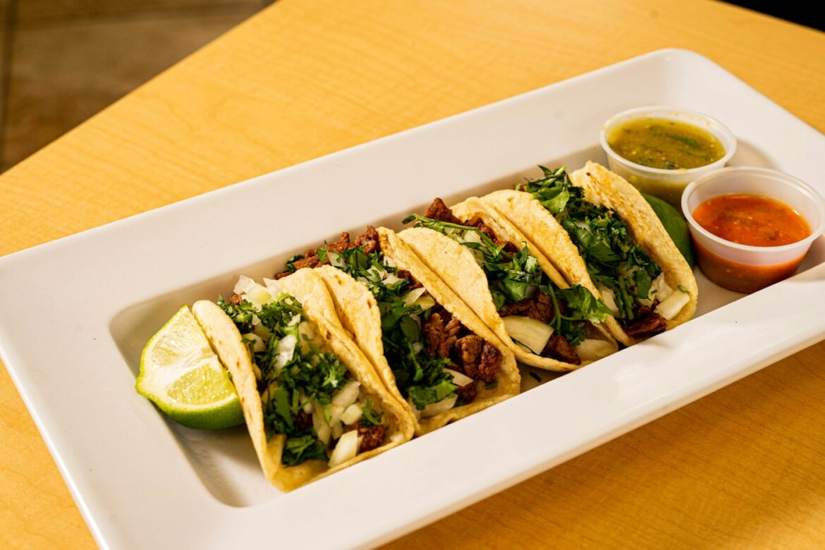 A white platter of soft corn tacos with cilantro and onion, served with lime and two salsas