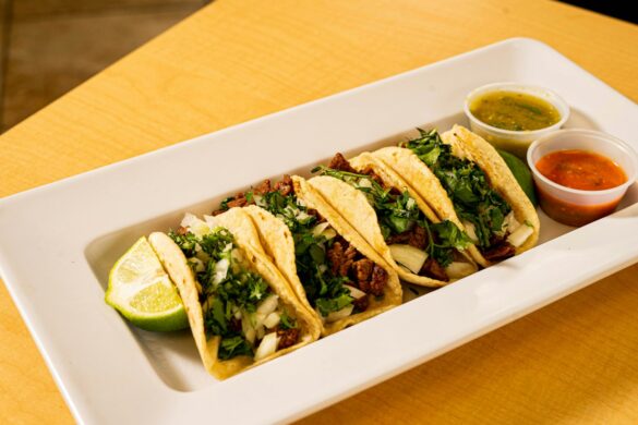 A white platter of soft corn tacos with cilantro and onion, served with lime and two salsas