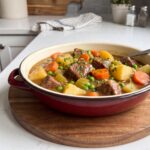 Crockpot beef stew with chunks of beef, potatoes, carrots, celery and peas in a thick gravy in a red enamel dish
